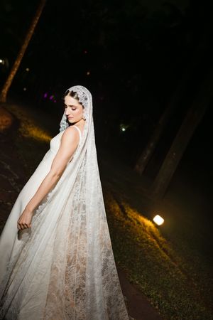 A bride in a flowing white lace veil poses outdoors at night with warm lighting in the background.