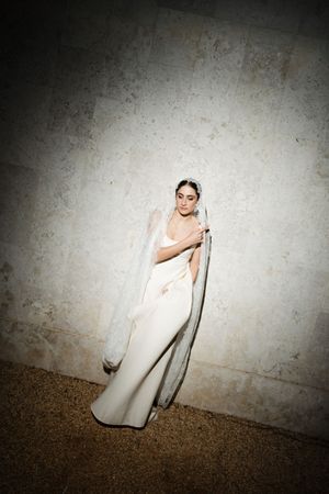 Someone in an elegant white wedding gown poses dramatically against a textured wall in moody lighting.