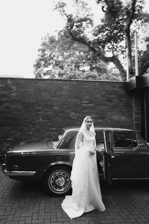 Classic black vintage car parked by brick wall with elegant wedding dress in black and white photography.