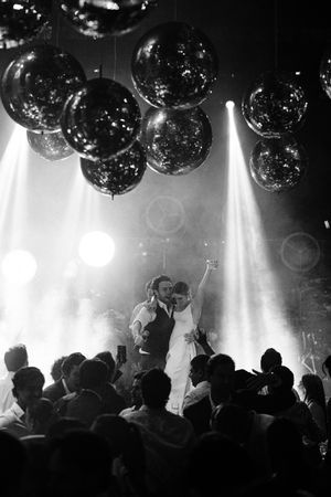 A black and white photo of a wedding celebration in a dark venue with large disco balls overhead and spotlights illuminating the crowd.