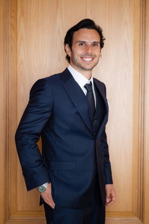 Professional portrait of a person in a dark navy suit and tie standing against a wooden paneled wall.