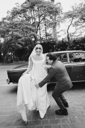 A black and white wedding photo showing a graceful bride in a flowing dress being helped on cobblestone street near vintage car.