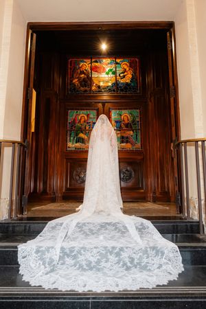 White wedding dress with dramatic cathedral length lace veil displayed on church steps in front of wooden doors.