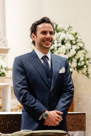 Groom wearing a navy suit smiles warmly during wedding ceremony with white floral arrangements in background.