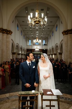 A couple celebrates their traditional church wedding ceremony inside an elegant chapel with crystal chandeliers and stone columns.