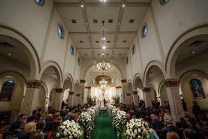 Interior view of a church wedding ceremony with arched ceilings, chandeliers and white floral arrangements lining the aisle.