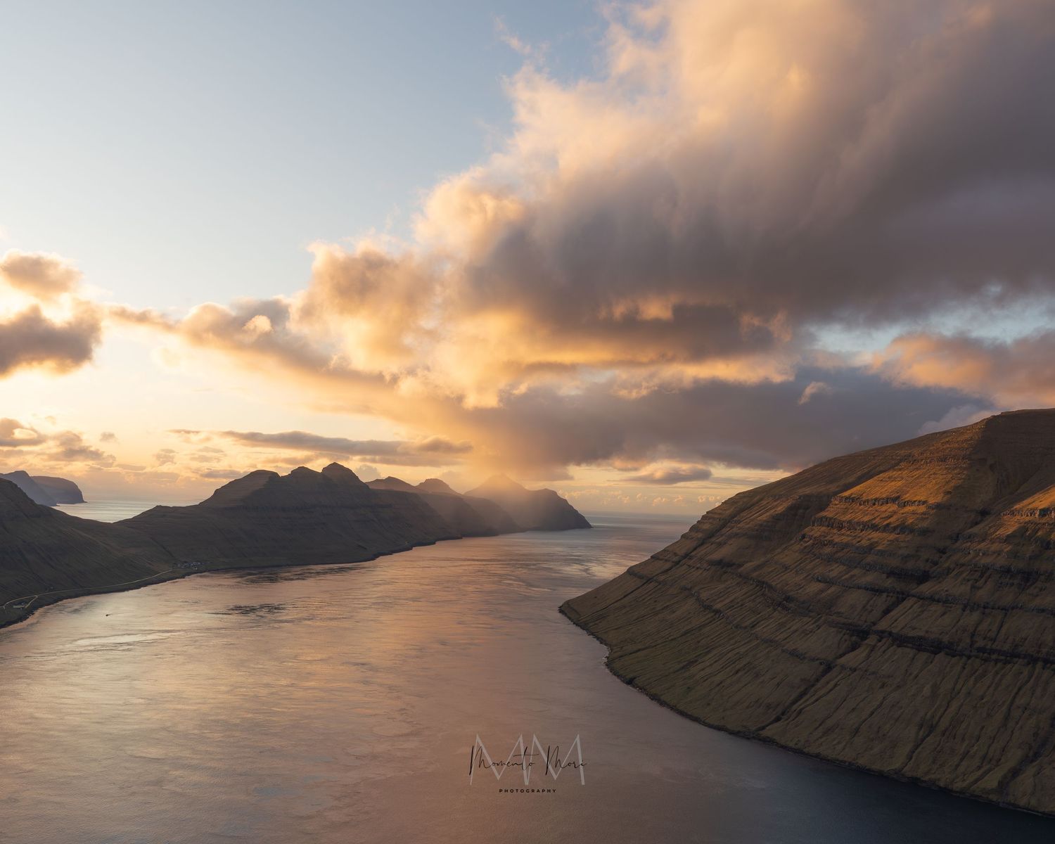 Pastel pink clouds hover over the fjords and the ocean in this landscape photograph taken at dusk in the Faroe Islands