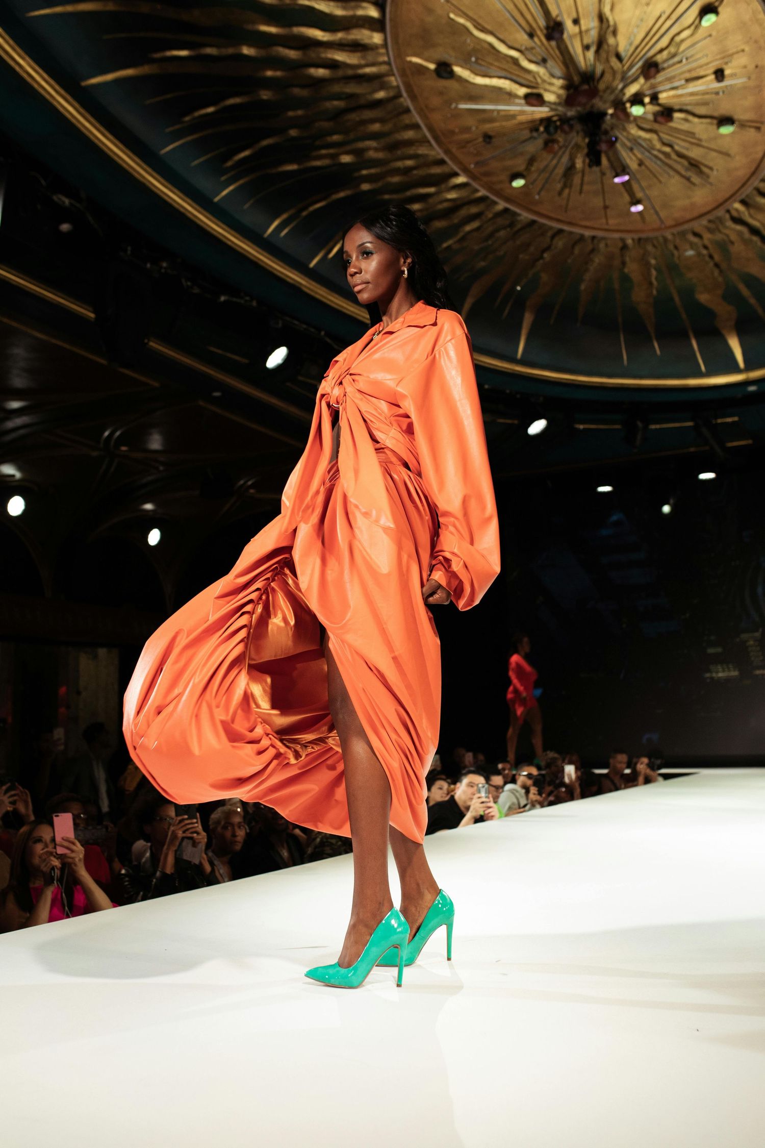 Fashion model wearing flowing orange dress and bright turquoise heels walks runway under ornate ceiling light fixture.