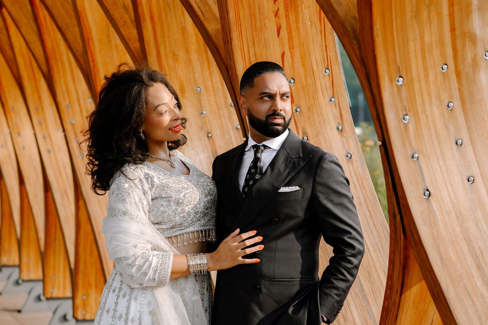 A couple in formal wedding attire poses in front of a wooden wall decorated with string lights and metallic accents.