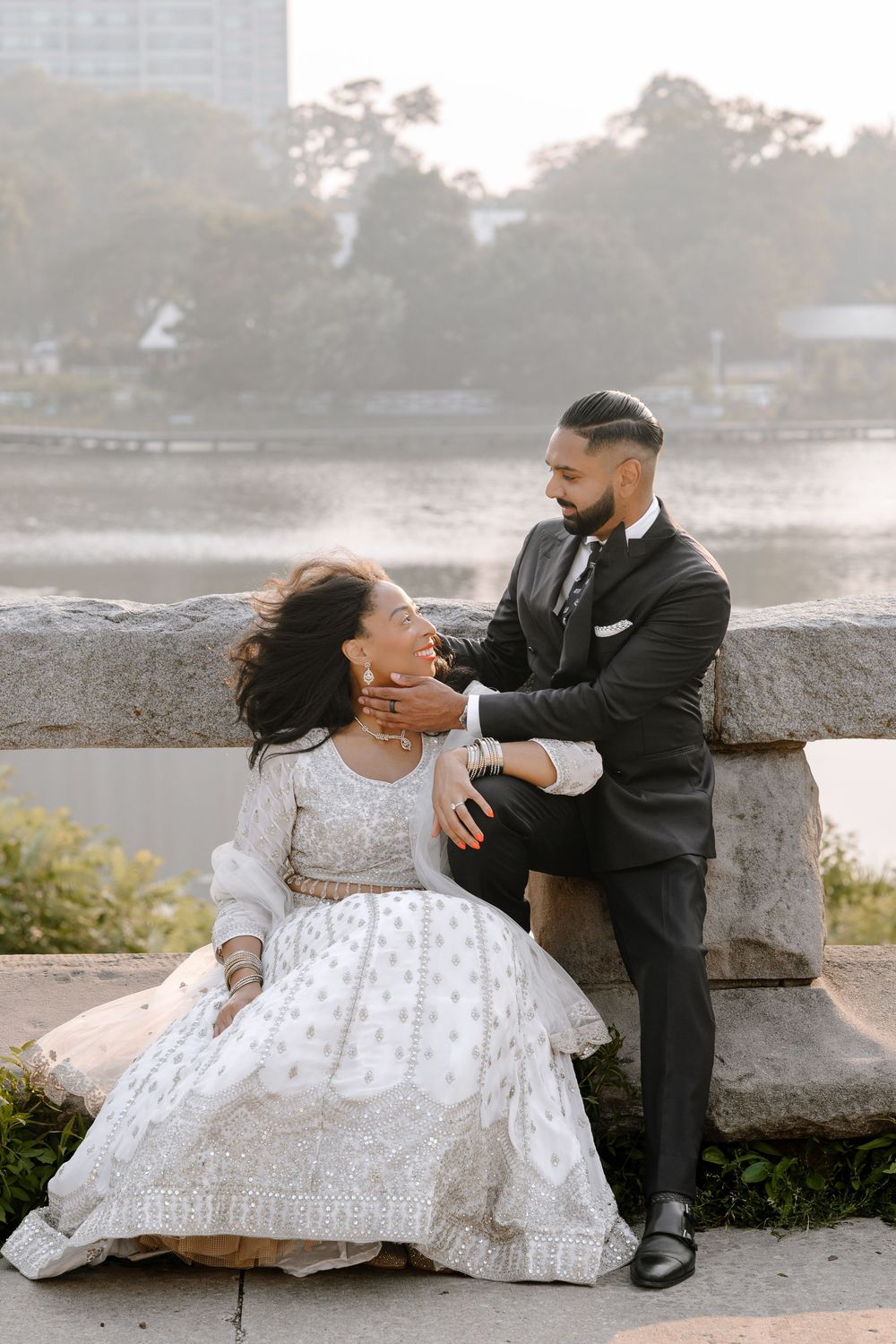 A couple shares a romantic moment by the water, with the bride wearing a flowing white lace gown and groom in black suit.