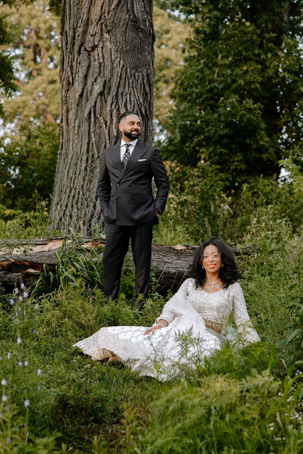 A couple poses for their wedding photos in a lush green outdoor setting with a large tree trunk backdrop.