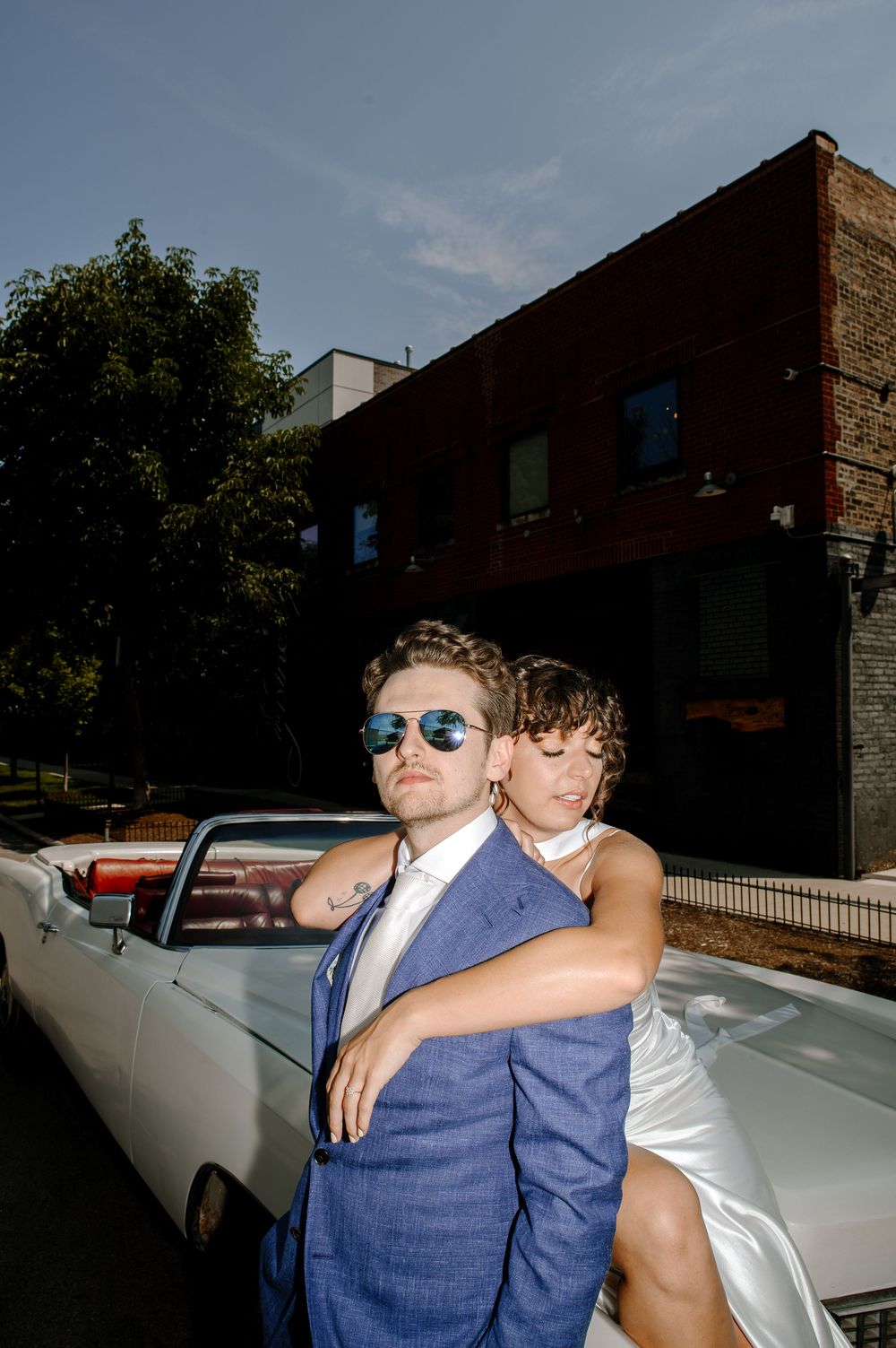 A couple poses with a vintage white convertible car outside a brick building on a sunny day.