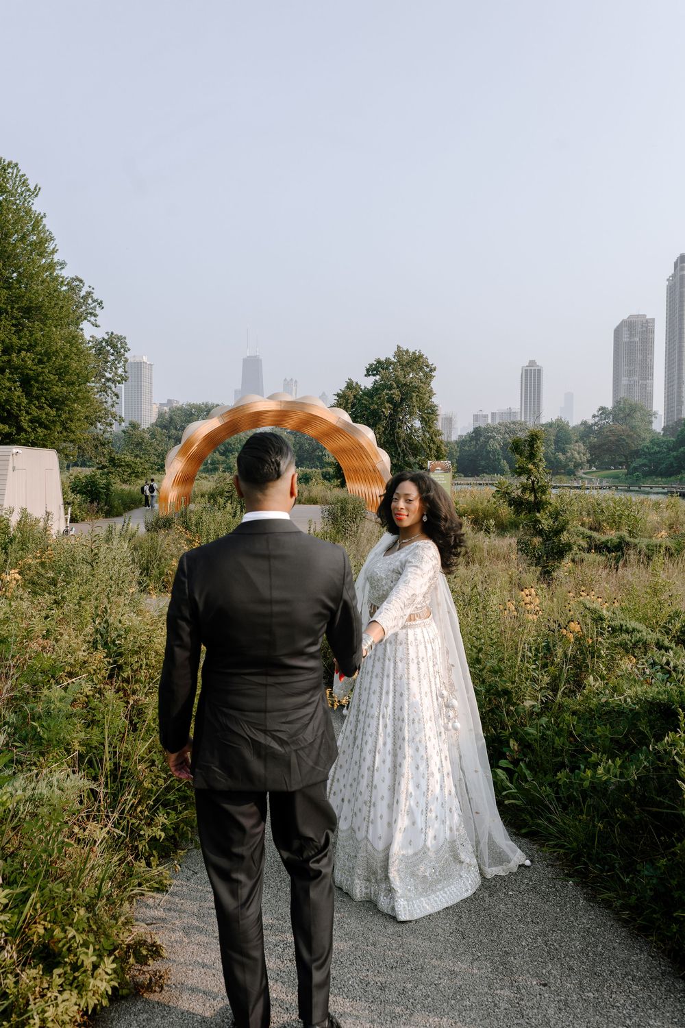 Couple in wedding attire standing on path in park with city skyline and orange smoke arch detail in background.
