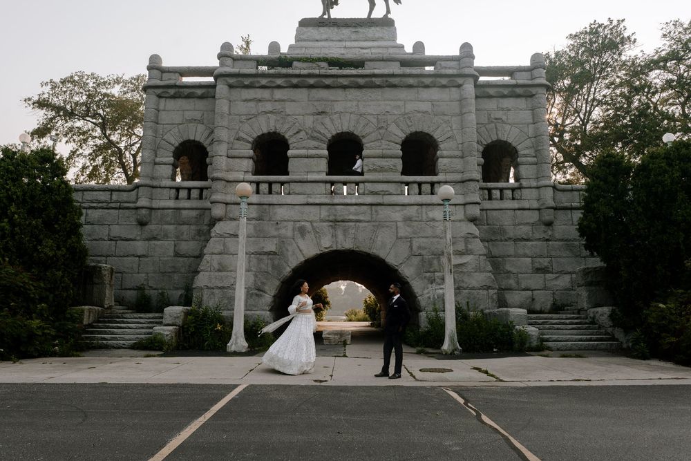 A wedding couple walks along a stone terrace with arches and steps overlooking greenery at a historic venue.