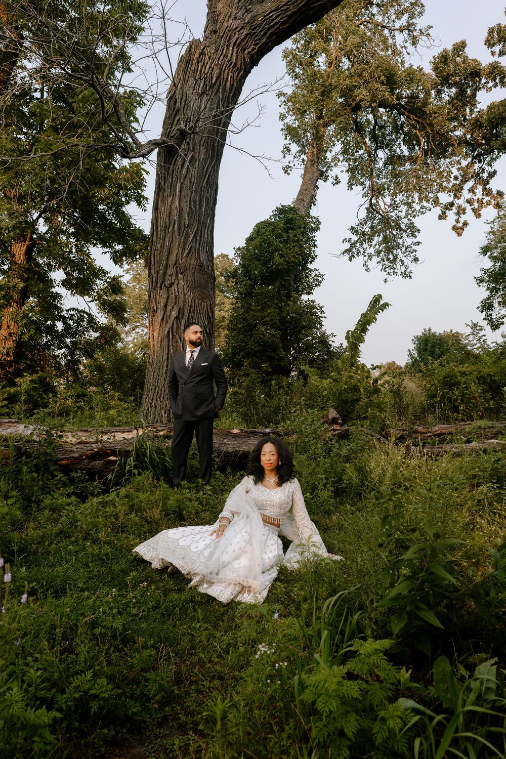 A couple poses in a scenic outdoor setting with lush greenery and a large tree trunk in golden hour lighting.