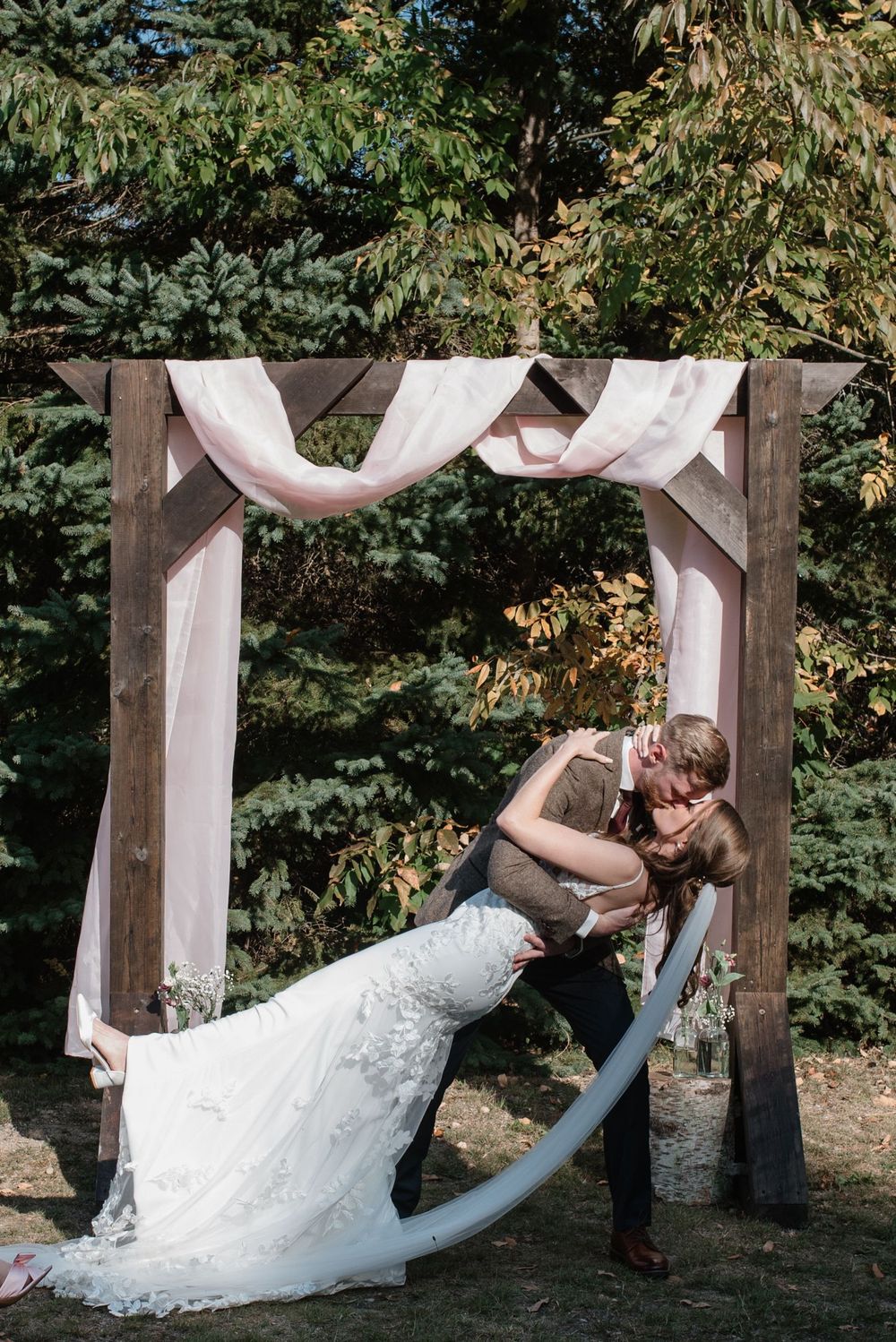 Couple in romantic pose under rustic wooden wedding arch draped with white fabric in outdoor forest setting.