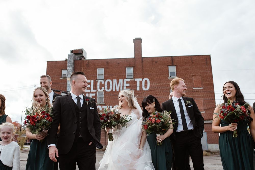 Wedding party walking and laughing together in front of brick wall with Welcome sign visible in background.
