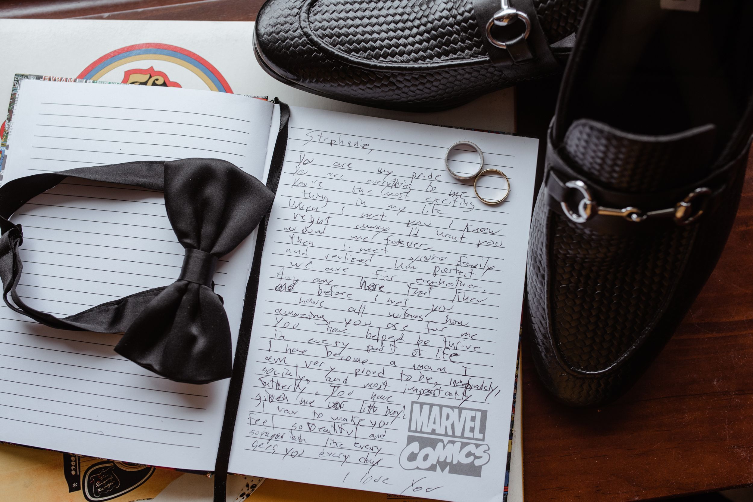 Black bow tie, wedding rings, and handwritten vows on lined paper next to dress shoes for wedding preparation.