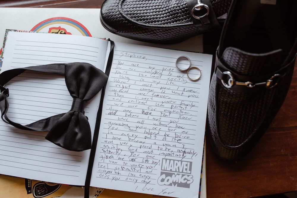 Black bow tie, wedding rings, and handwritten vows on lined paper next to dress shoes for wedding preparation.