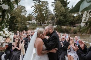 Happy couple kissing at outdoor wedding ceremony while guests celebrate and cheer in garden setting.