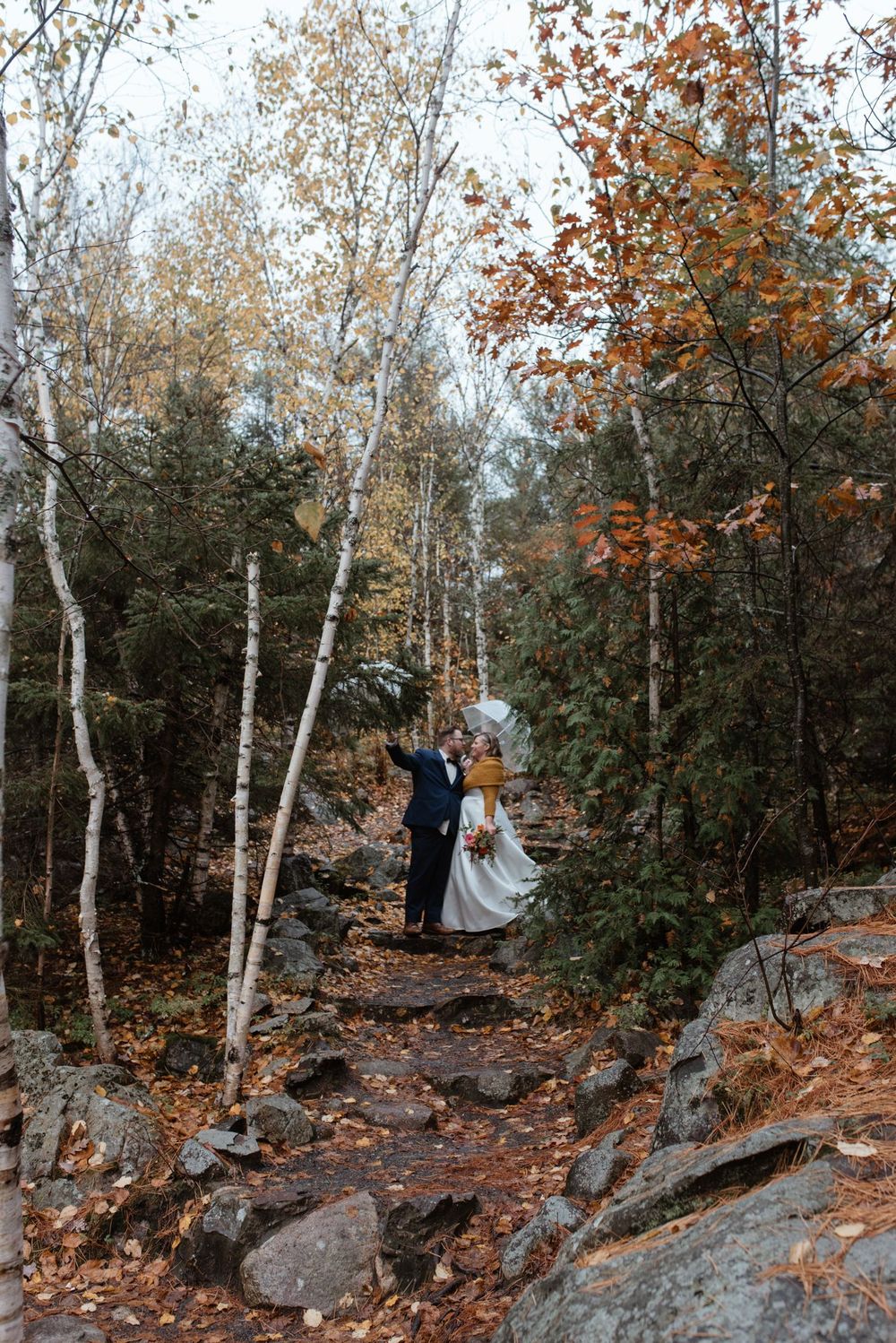 Couple standing on a rocky autumn forest trail surrounded by birch trees and orange fall foliage.