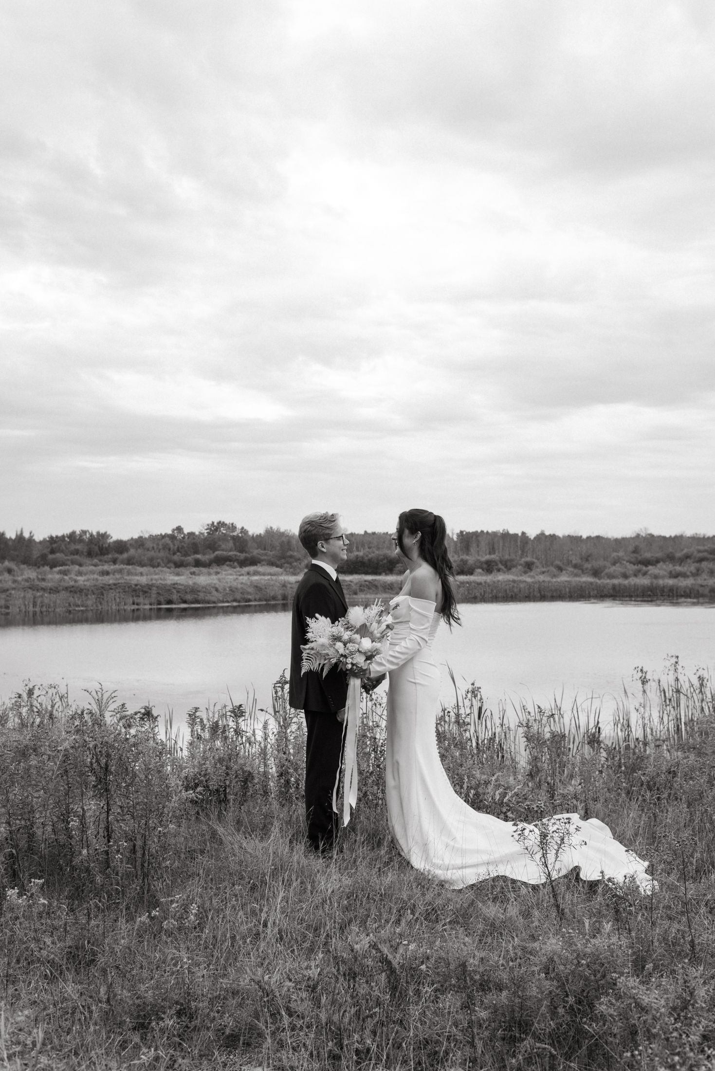 A romantic black and white wedding photograph of a couple standing by a serene lake surrounded by tall grasses.