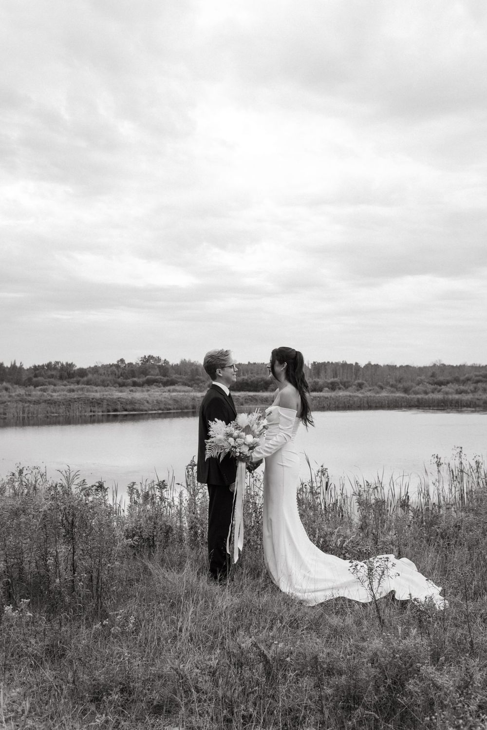 A romantic black and white wedding photograph of a couple standing by a serene lake surrounded by tall grasses.