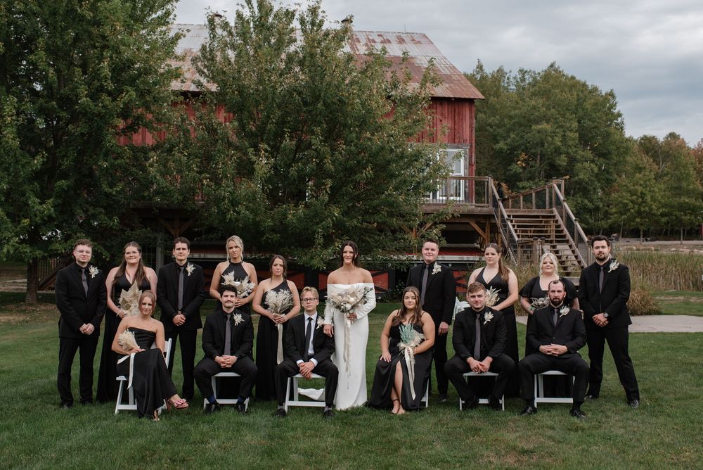 A wedding party stands on green grass in front of a rustic red barn on a cloudy autumn day.