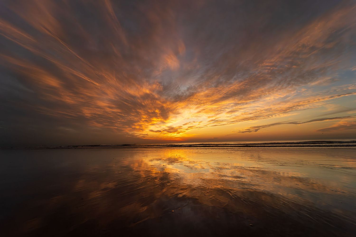 Dramatic orange and gold sunrise reflects perfectly in calm ocean shoreline creating mirror-like symmetry with fiery clouds above.
