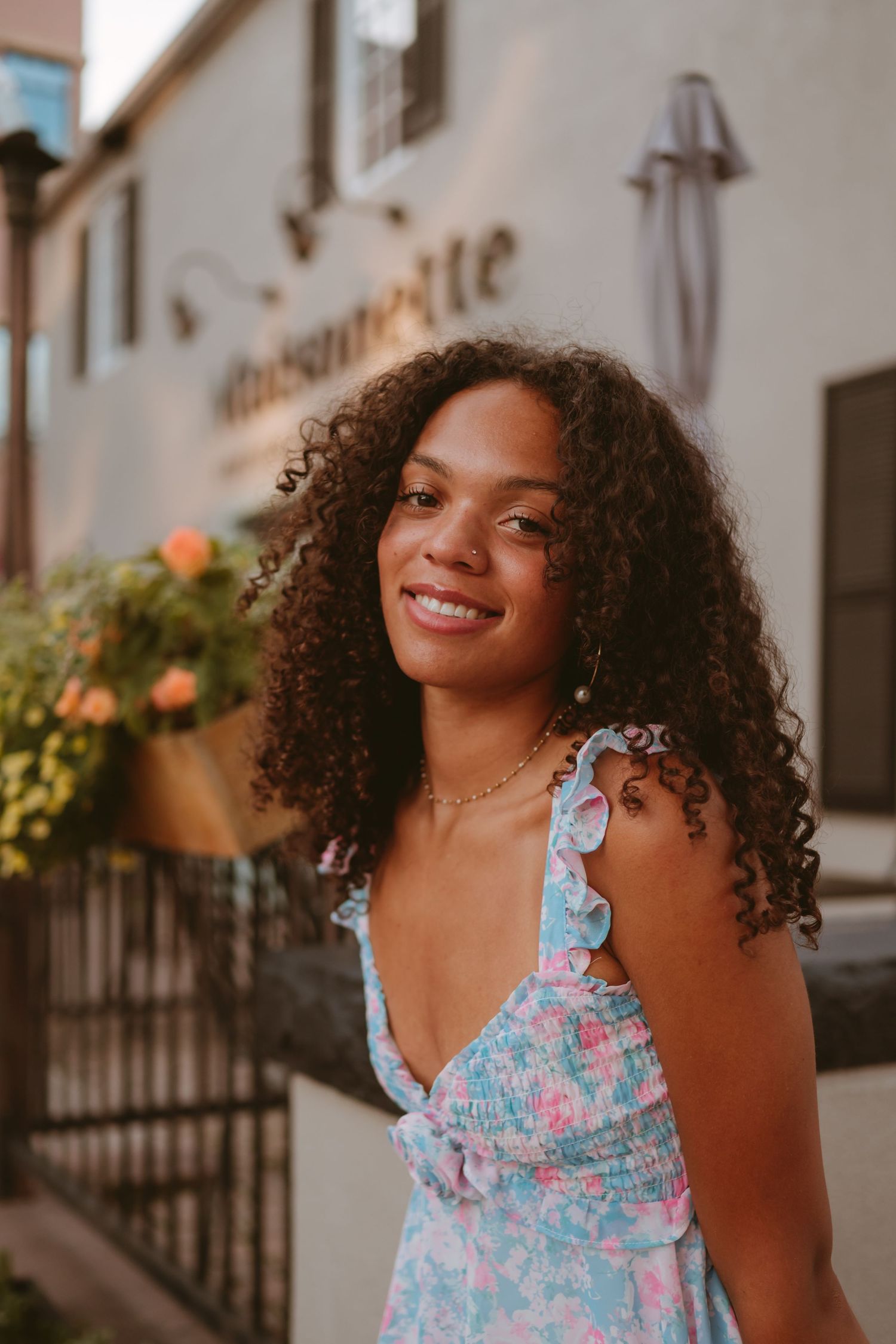 Individual wearing a floral ruffled sundress smiles warmly in an outdoor setting with soft evening light.