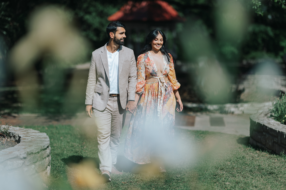 A couple walks together in a garden setting, wearing elegant wedding attire with dreamy bokeh effects in the foreground.