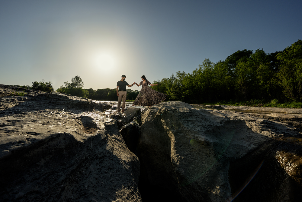 Silhouettes of people hiking on rocky terrain against bright sunlit sky and green trees in background.