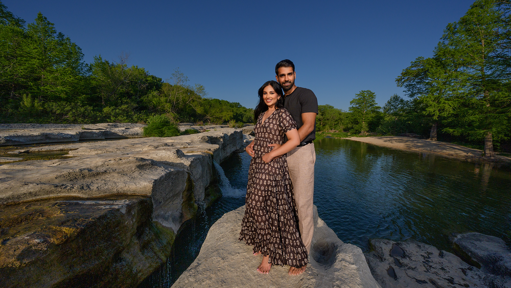 A couple embraces while standing on rocky outcrops near a serene pond surrounded by green trees on a sunny day.