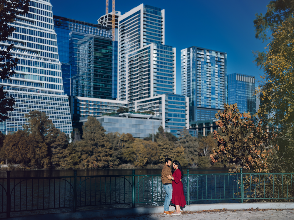 A couple embraces along Lady Bird Lake with Austin's modern blue skyscrapers reflecting in the water on a sunny day.
