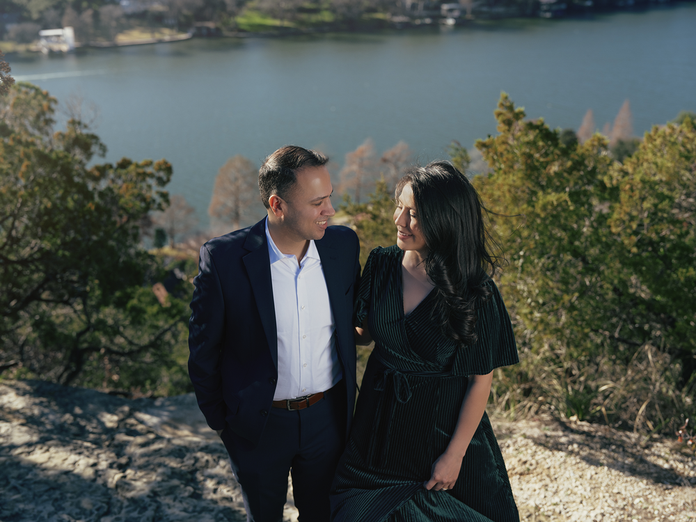 A couple in dark clothing stands on a scenic overlook above a calm lake on a sunny day.