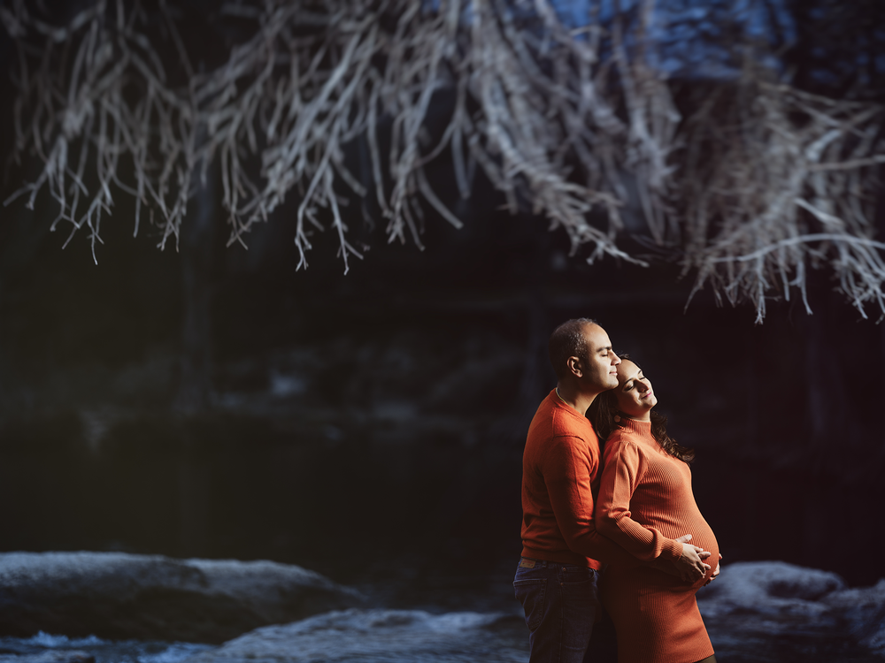 Two people in bright orange jackets embrace under frosty winter branches near a dark body of water.