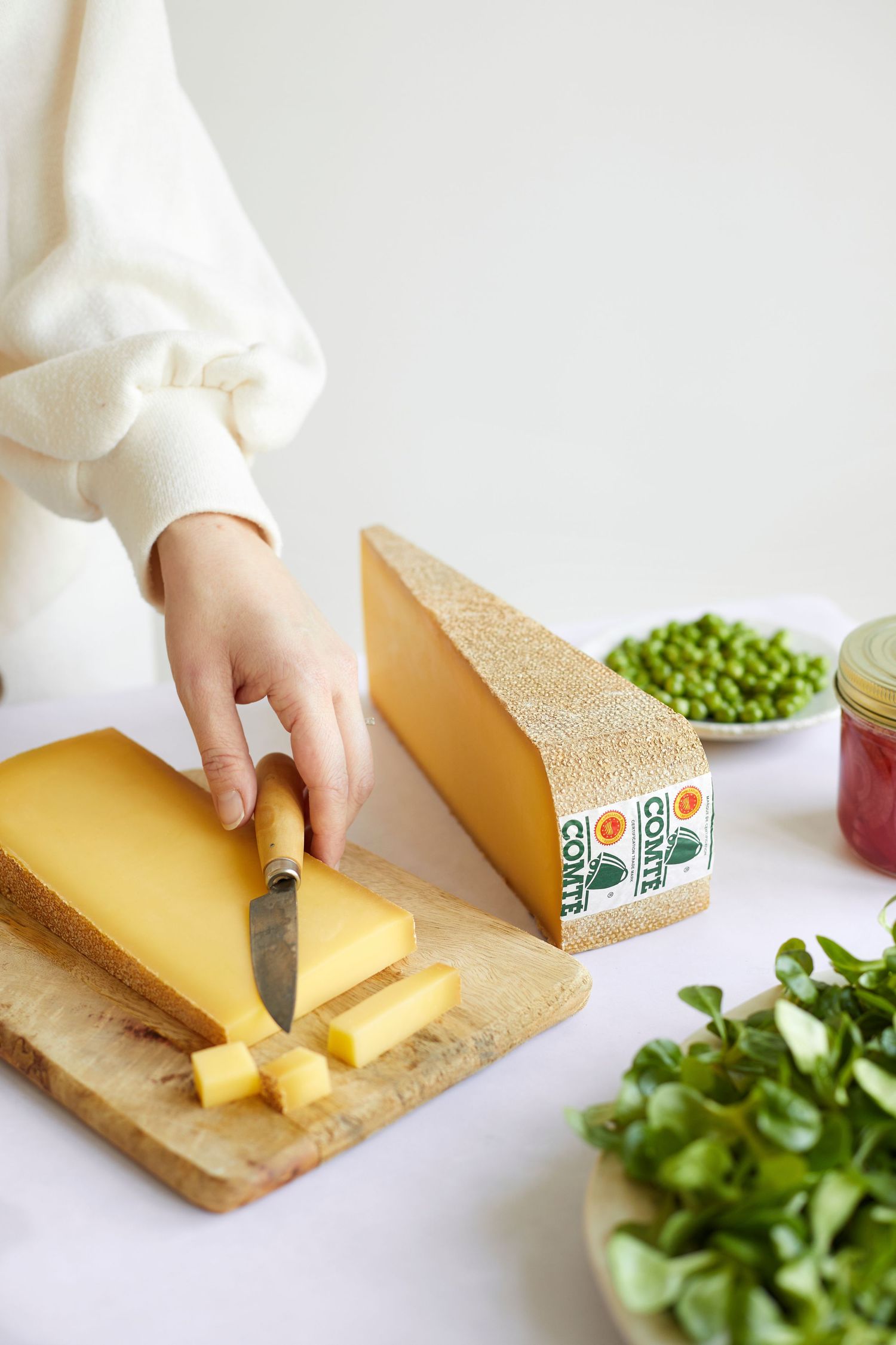 Someone slices a large block of yellow cheese on a wooden cutting board with fresh greens and peas visible nearby.