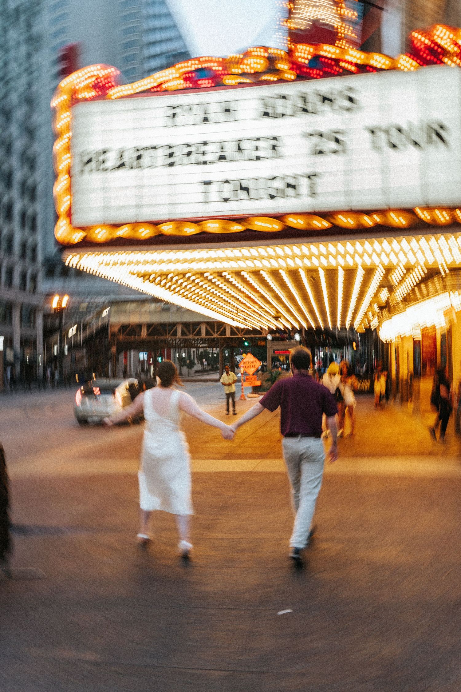 Motion blur captures people walking under an illuminated theater marquee at night, creating a dynamic urban scene.