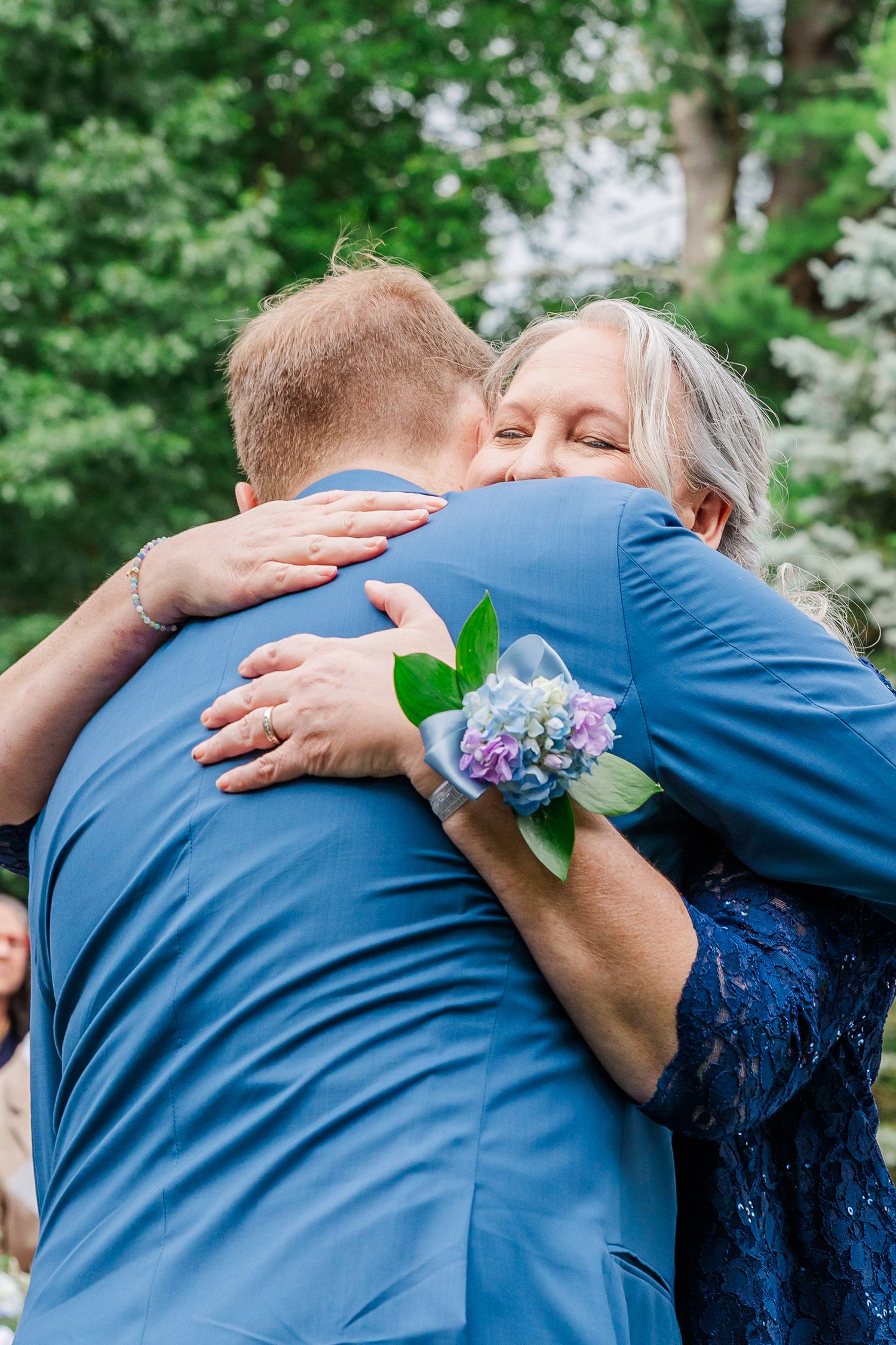 Two people share an emotional embrace at an outdoor event, with a blue hydrangea corsage and green foliage in the background.