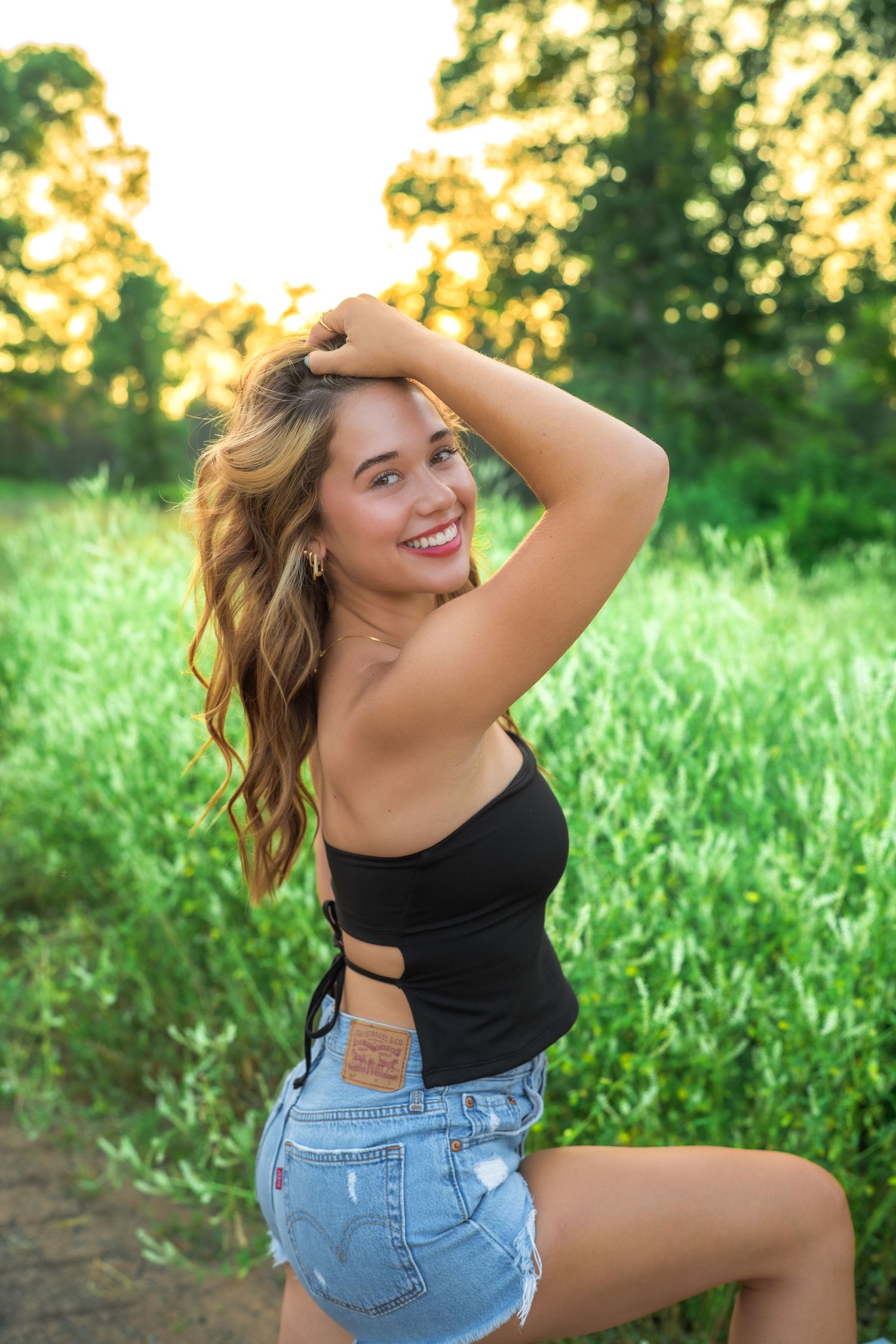 Carefree person in black crop top and denim shorts poses outdoors during golden hour in lush green field.