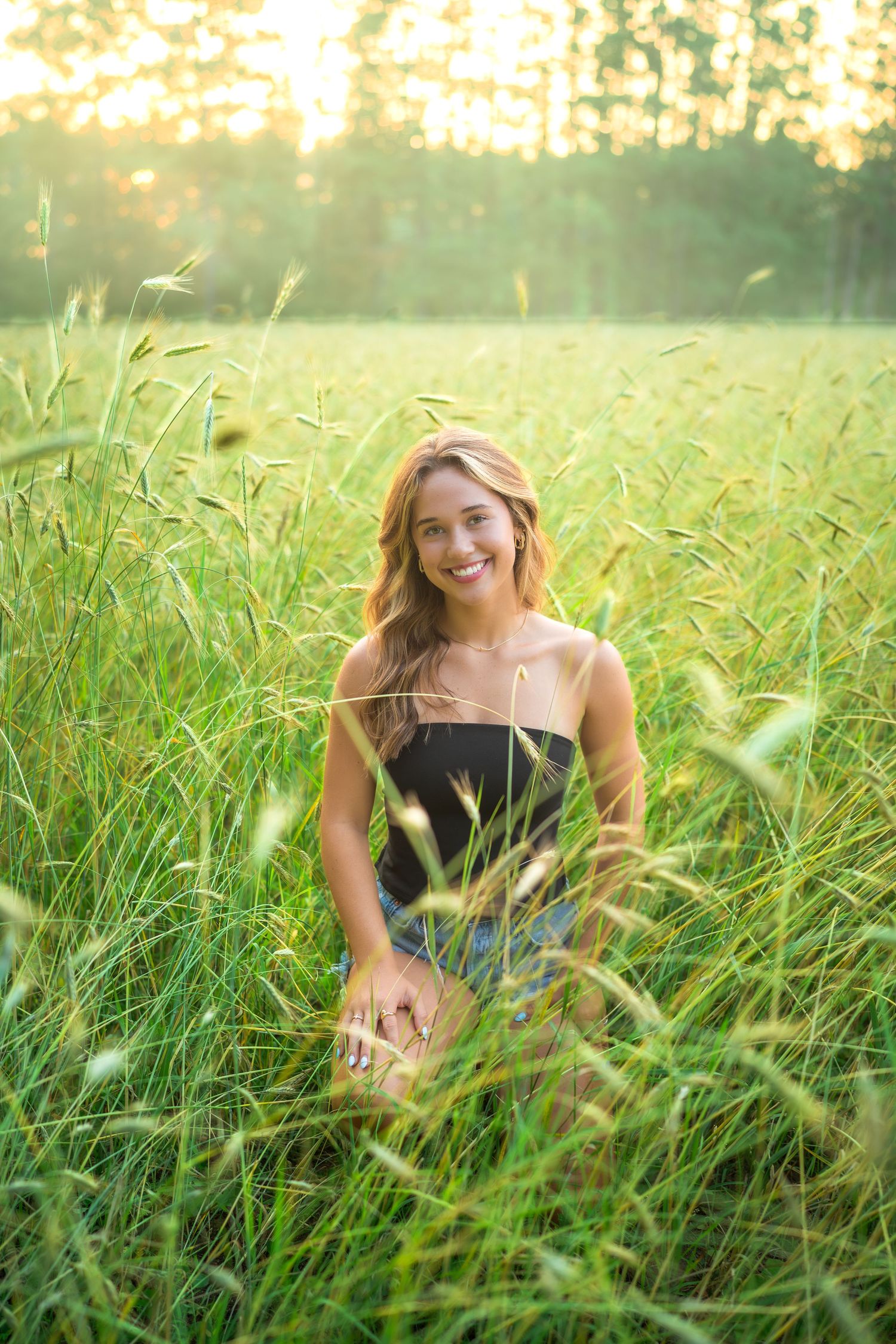 A joyous woman in a black dress sits among tall grass at golden hour with sunlit trees in the background.