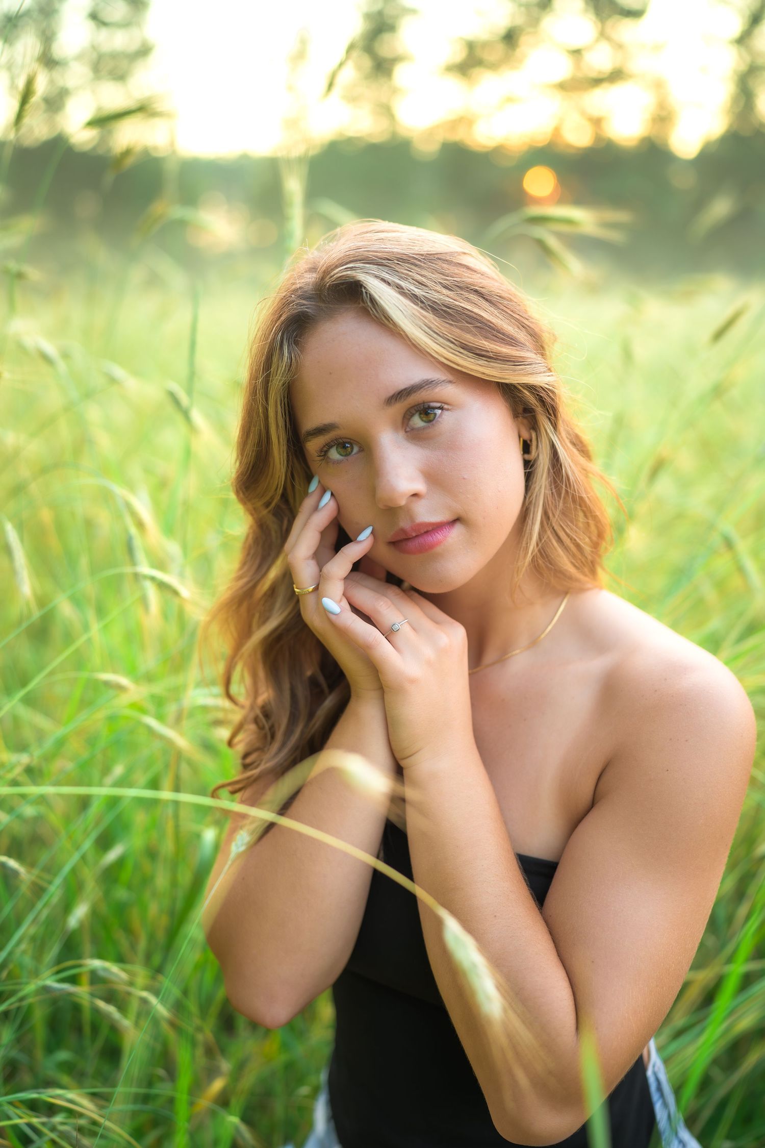 Young woman in black top poses gracefully near tall grass during a golden hour portrait photoshoot.