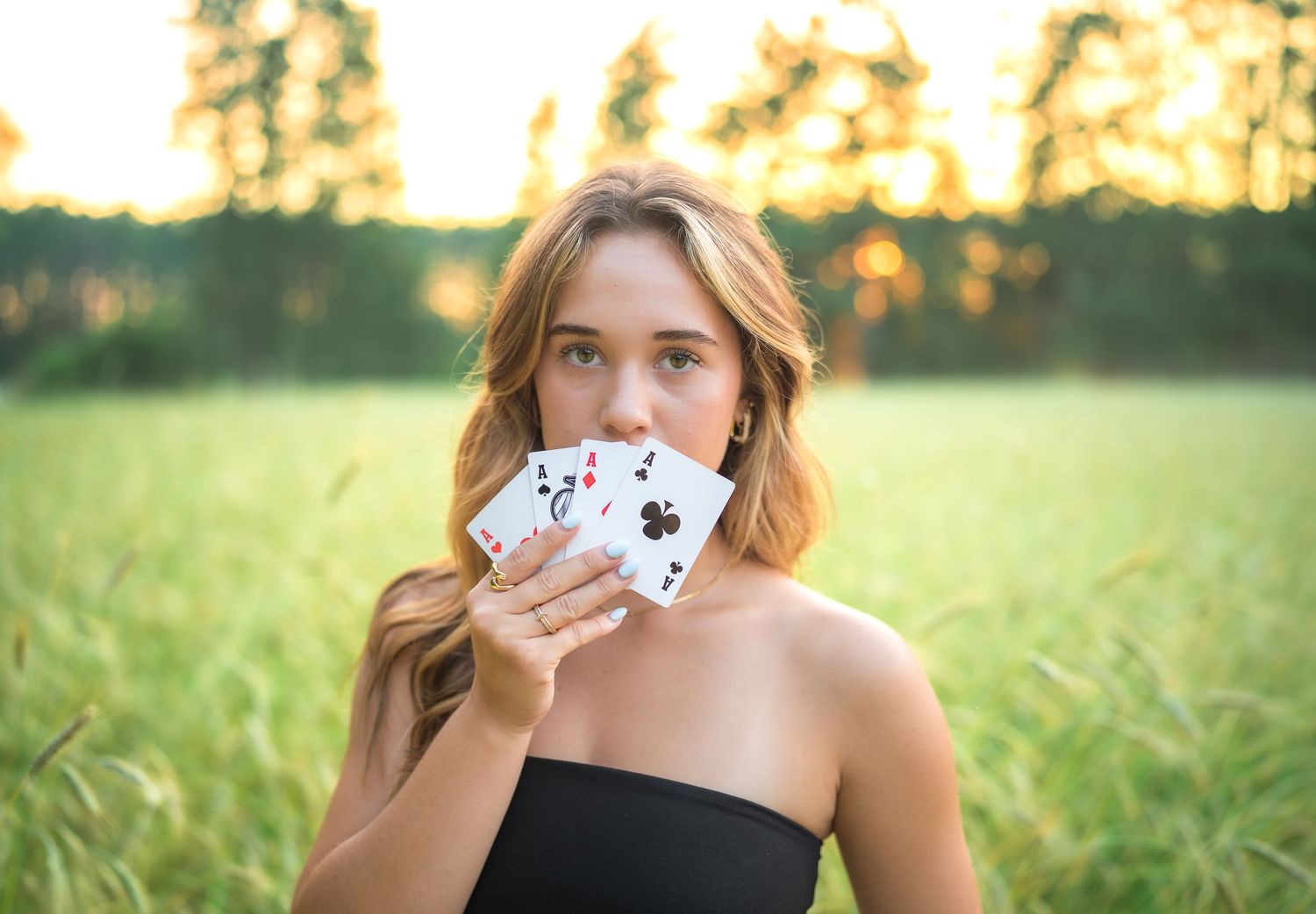 Person holding playing cards in front of face while posing in a grassy field at golden hour sunset.