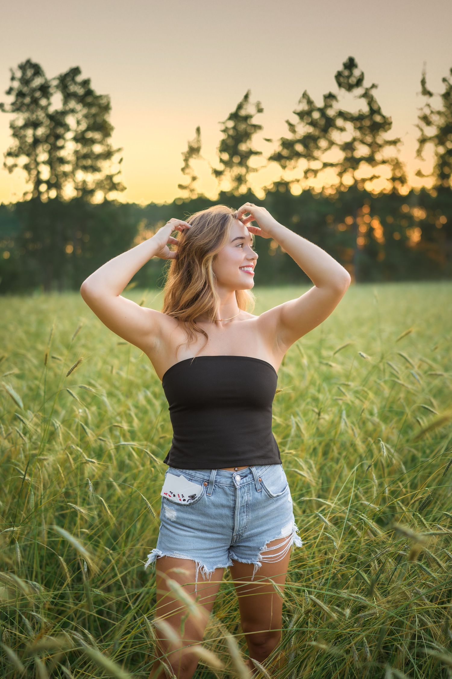Woman in denim shorts and black top poses in golden wheat field at sunset with trees in background.
