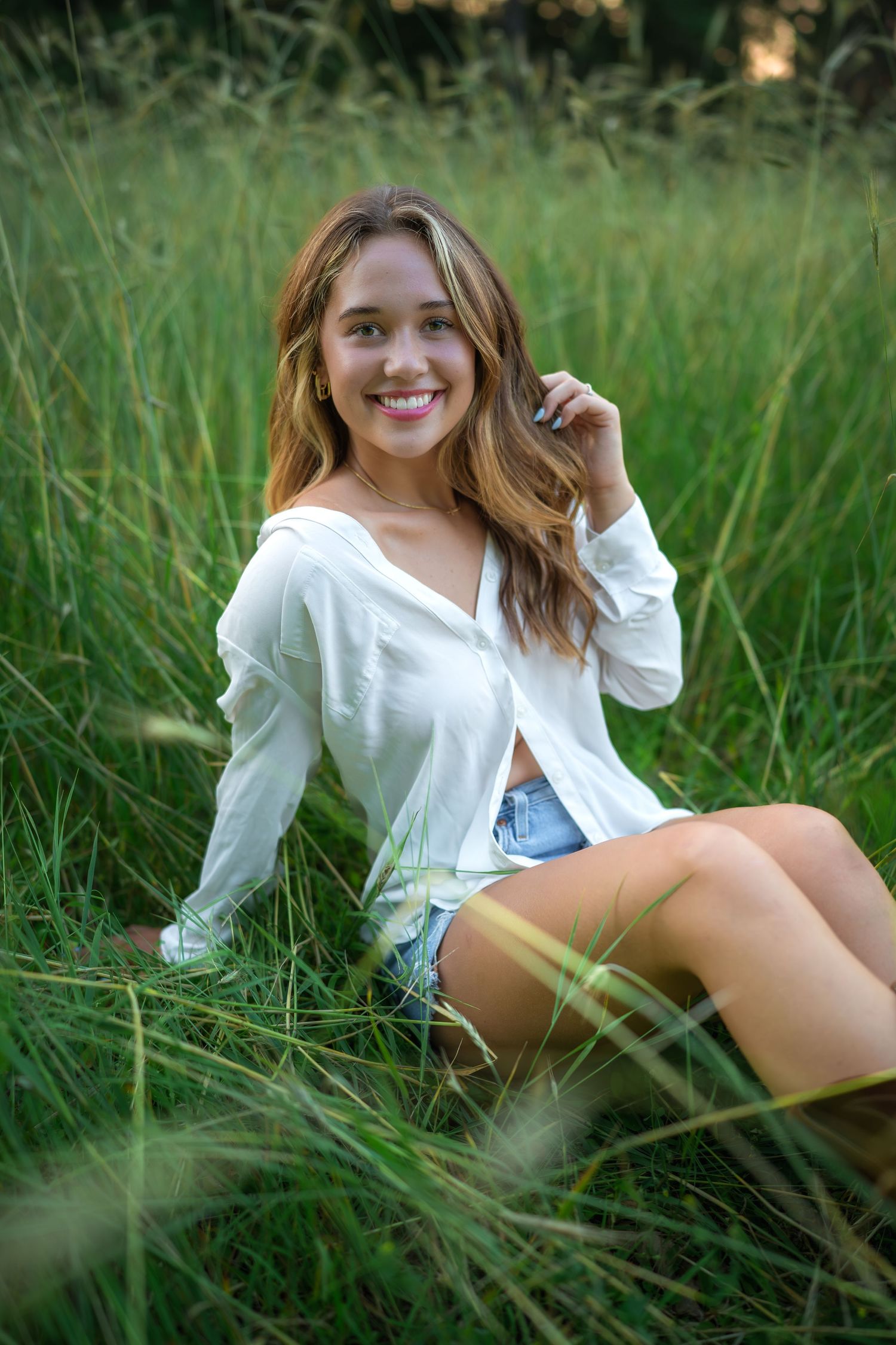 A candid outdoor portrait in tall green grass featuring a white button-down top and denim shorts on a summer day.