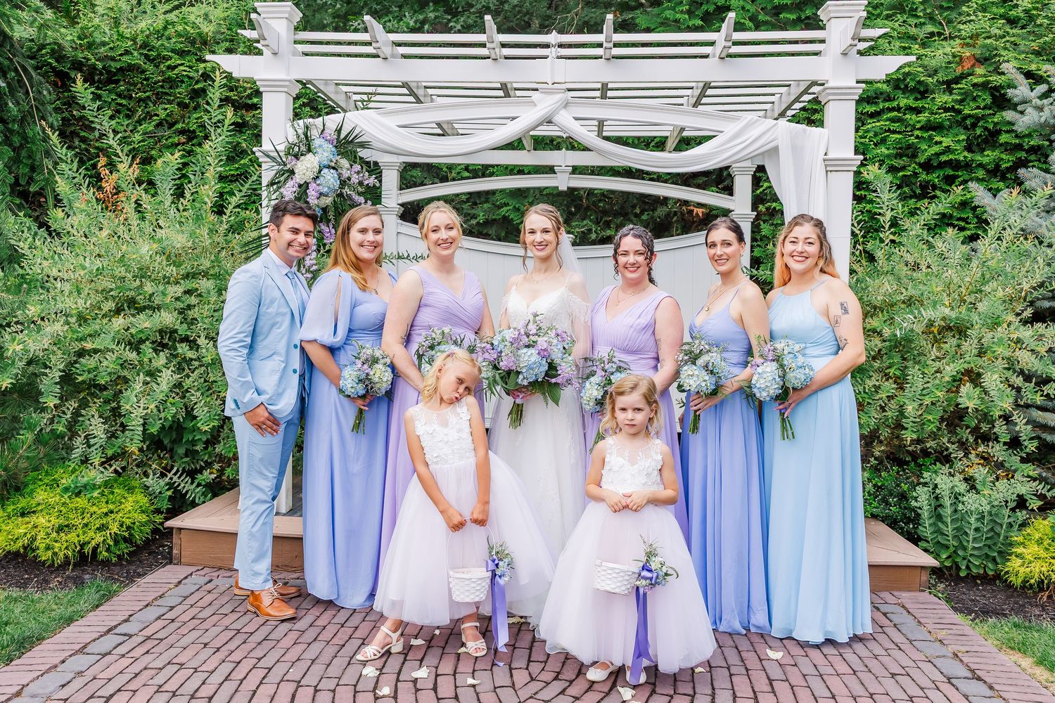 A wedding party in lavender and blue dresses poses together under a white garden arbor during an outdoor ceremony.