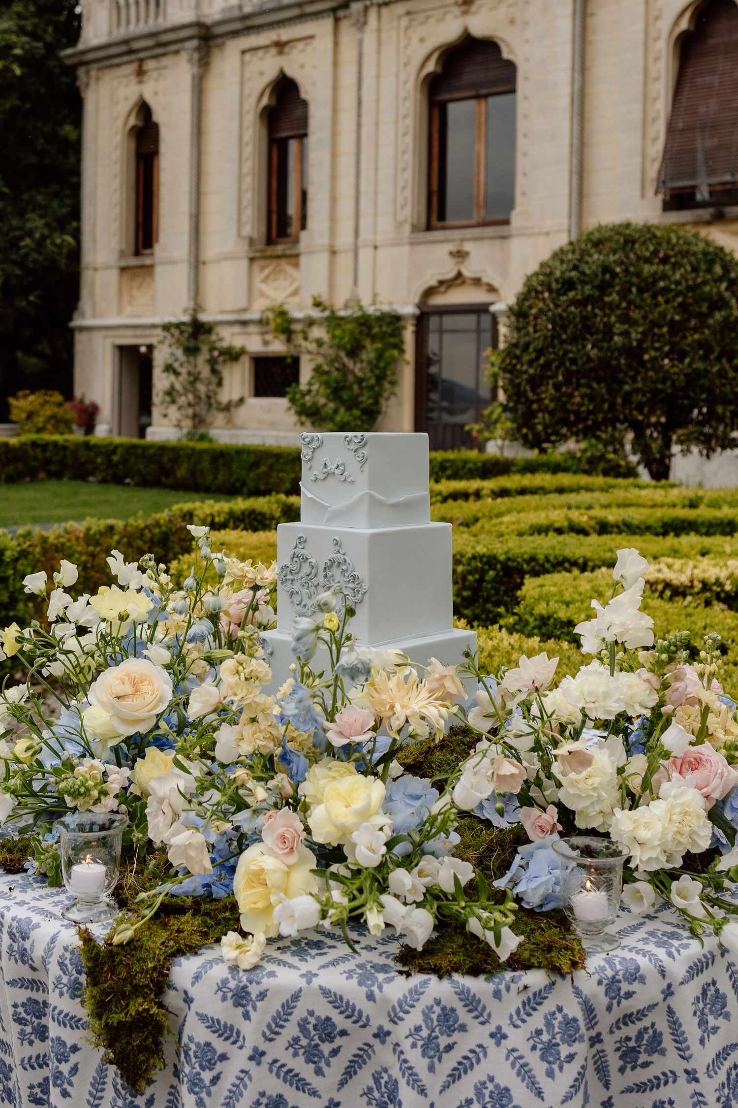 Wedding cake and floral centerpieces in pastel blue and pink displayed on an ornate table with mansion gardens backdrop.