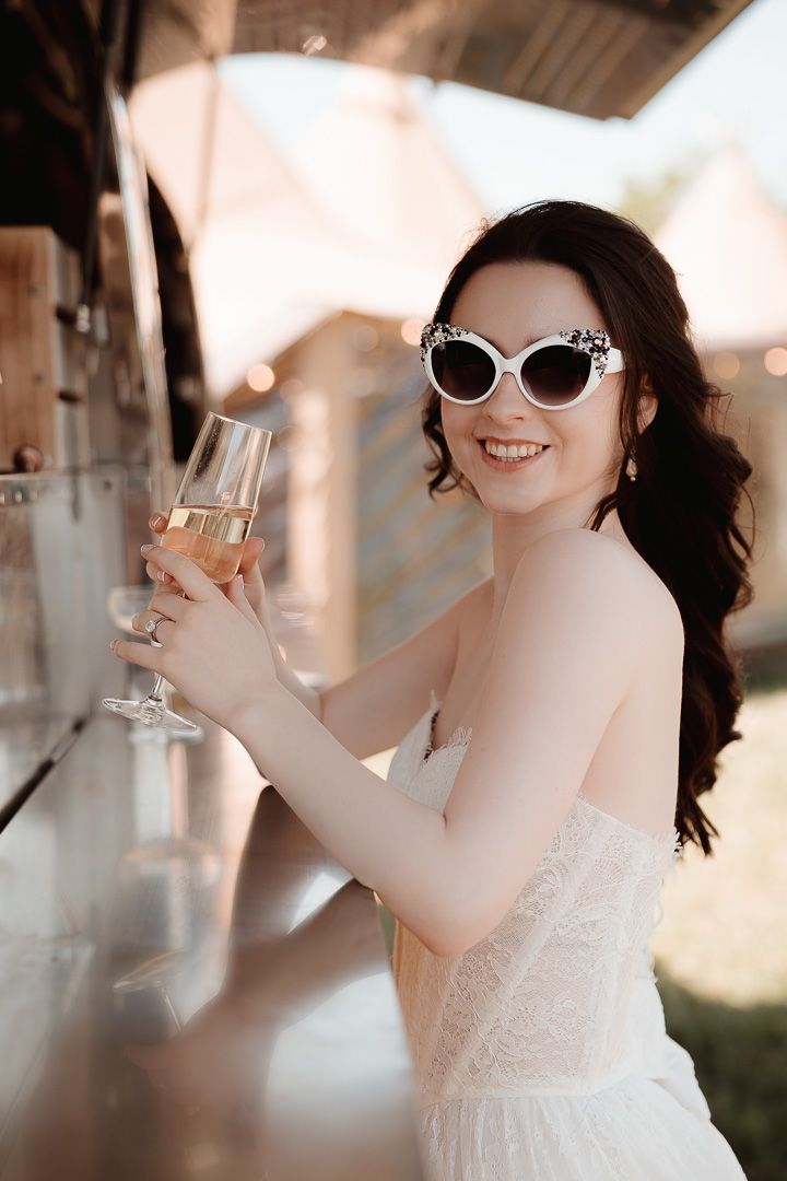 A joyful figure in white lace dress and stylish sunglasses holds up a champagne glass with an elegant smile.