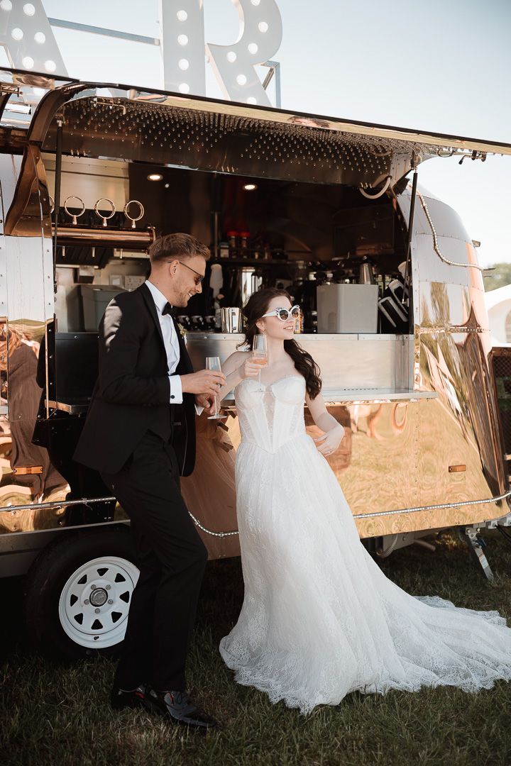 A couple stands beside an elegant vintage mobile bar trailer at an outdoor wedding event during golden hour.