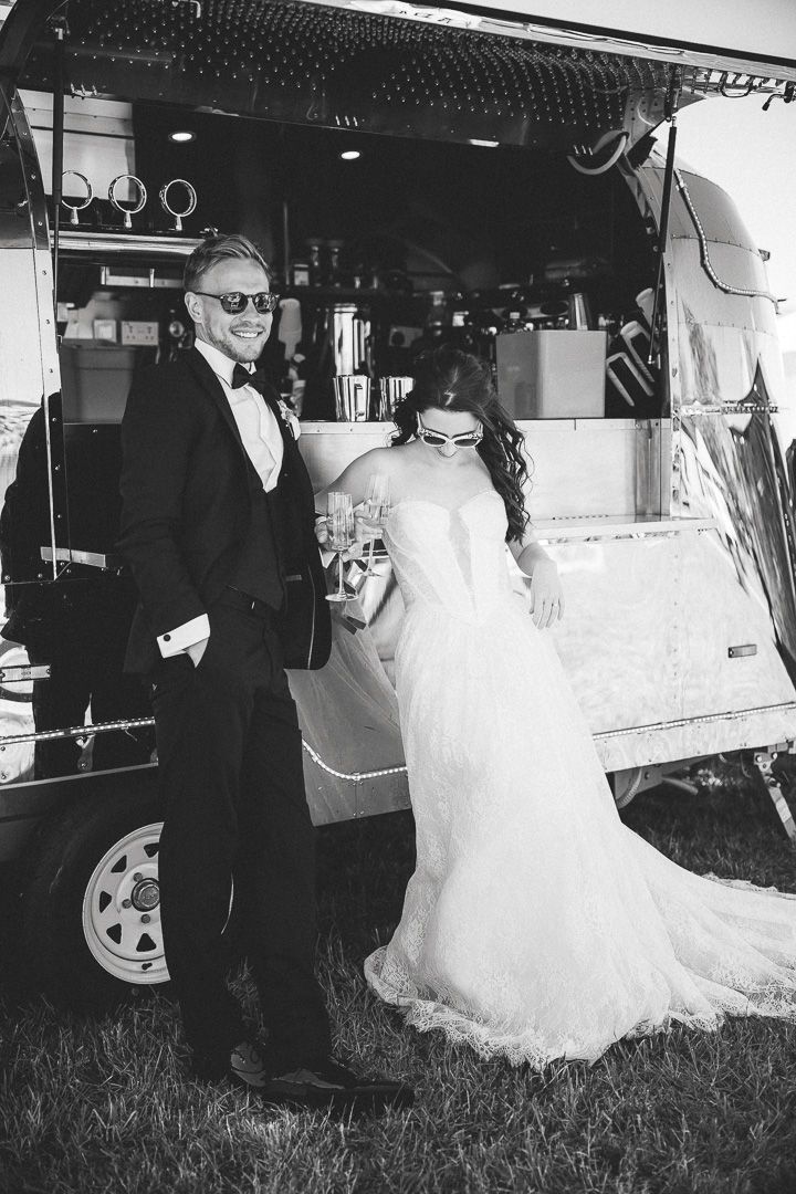 Black and white wedding photo with couple posing next to mobile beverage truck outdoors on grass.